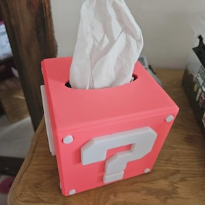 Peut inclure: Boîte à mouchoirs rose en forme de bloc point d'interrogation d'un jeu vidéo. Le bloc présente un point d'interrogation blanc et des détails blancs. Des mouchoirs blancs dépassent du haut. Posé sur une surface en bois.
