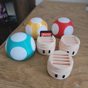 Peut inclure: Ensemble de supports de cartouches de jeu et de boules décoratives en forme de champignon, imprimés en 3D. Les supports sont rose clair avec des yeux noirs et une fente pour les cartouches de jeu. Les boules sont bleues, jaunes et rouges avec des pois blancs.