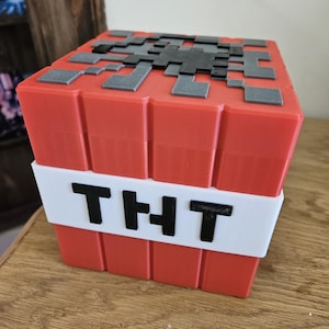 Può includere: Un blocco TNT di Minecraft rosso e bianco con un'etichetta 'TNT' nera. Il blocco è fatto di plastica e ha un design a pixel.