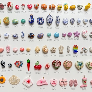 K&ouml;nnte beinhalten: Eine Sammlung kleiner Anh&auml;nger aus Keramik und Harz, die in Reihen angeordnet sind und verschiedene Formen wie Tiere, Blumen, Herzen, Lebensmittel und Symbole aufweisen, jeweils mit einem eindeutigen alphanumerischen Code zur Identifizierung.