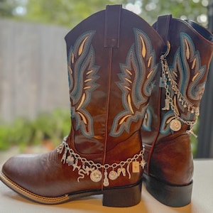 May include: A pair of brown leather cowboy boots featuring intricate blue and cream embroidery on the shaft. The boots are adorned with silver-toned metal charm chains, including various small pendants like a guitar, clover, and star.
