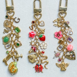 Puede incluir: Tres llaveros de metal dorado con varios dijes, incluyendo letras, formas de esmalte de colores, piedras preciosas y objetos en miniatura como una mariquita, un avión, una bota y una concha, unidos a un mosquetón y una cadena de eslabones.