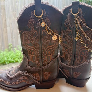 Puede incluir: Un par de botas vaqueras de cuero marrón oscuro con bordados en tono canela, bordes deshilachados y dijes de metal dorado sujetos a las trabillas. Los dijes incluyen cadenas, pequeñas cuentas, un ojo turco y un colgante de estrella.