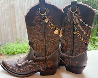 Può includere: Un paio di stivali da cowboy in pelle marrone scuro con ricami color cuoio, bordi sfrangiati e ciondoli in metallo dorato attaccati alle linguette. I ciondoli includono catenelle, piccole perline, un occhio e un pendente a stella.