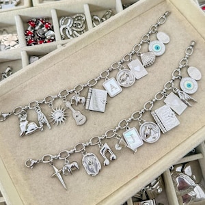 Puede incluir: Dos pulseras de dijes plateadas con una variedad de colgantes de metal, que incluyen un gato, sol, guitarra, elefante, libro, letra A, letra J, cartas de juego, un ojo turco, una cruz, una rosa, una nota musical y una copa de cóctel.