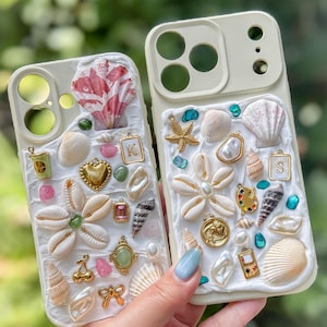May include: Two cream-coloured phone cases decorated with a textured white base layer, various seashells, gold-toned charms, and colourful resin accents. One case features a letter K charm, while the other displays a letter S charm.