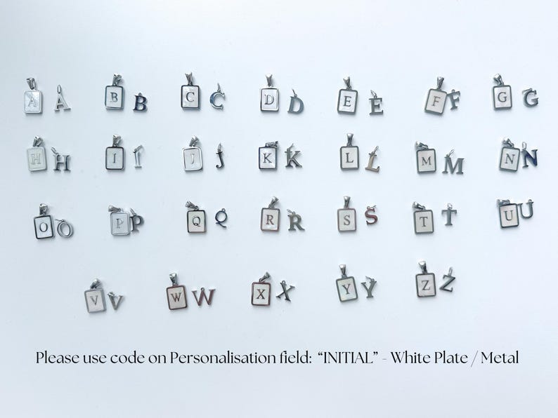 K&ouml;nnte beinhalten: Eine Sammlung von silberfarbenen Metall-Initialen-Anh&auml;ngern, jeweils mit einer rechteckigen wei&szlig;en Platte mit schwarzem Buchstaben oder einem einzelnen Metallbuchstaben-Anh&auml;nger. Der Text unten lautet: Please use code on Personalisation field: INITIAL - White Plate / Metal.