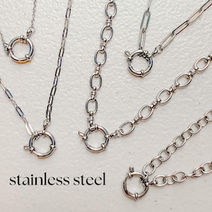 Puede incluir: Una colección de collares de acero inoxidable en tono plateado con varios estilos de eslabones, incluyendo eslabones ovalados alargados y eslabones redondos, cada uno asegurado con un broche de resorte circular. El texto stainless steel es visible.