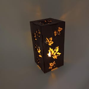 May include: A wooden lamp with a rectangular shape and cut-out maple leaf designs. The lamp is lit with a warm yellow light.