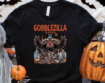 Könnte beinhalten: Schwarzes T-Shirt mit einem Halloween-Motiv. Das Design zeigt einen Truthahn mit ausgebreiteten Flügeln, den Text "GOBBLEZILLA" in Orange und japanische Schriftzeichen. Das T-Shirt ist von Halloween-Dekorationen umgeben, darunter Kürbisse, Spinnen und Fledermäuse.