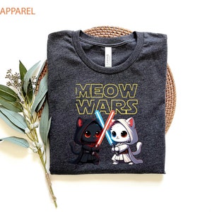 May include: A dark gray t-shirt with the text "MEOW WARS" in yellow. Two cartoon cats are holding lightsabers, one black cat with a red lightsaber and one white cat with a blue lightsaber.