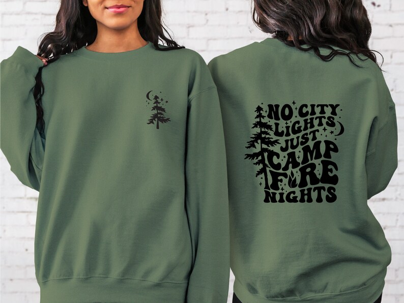 K&ouml;nnte beinhalten: Ein gr&uuml;ner Pullover mit einem schwarzen Design auf der R&uuml;ckseite, das "No City Lights Just Camp Fire Nights" sagt. Die Vorderseite des Pullovers hat ein kleines schwarzes Baumdesign.