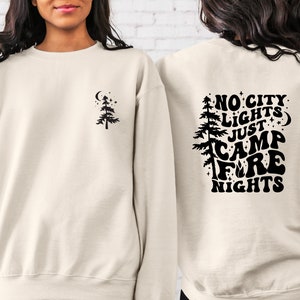 K&ouml;nnte beinhalten: Ein cremefarbener Pullover mit einem schwarzen Design auf der R&uuml;ckseite, das "No City Lights Just Camp Fire Nights" mit einem Baum und Sternen zeigt. Die Vorderseite des Pullovers hat ein kleines schwarzes Baumdesign.
