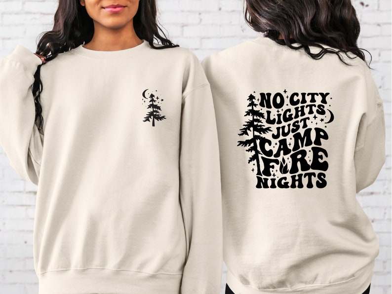 K&ouml;nnte beinhalten: Ein cremefarbener Pullover mit einem schwarzen Design auf der R&uuml;ckseite, das "No City Lights Just Camp Fire Nights" mit einem Baum und Sternen zeigt. Die Vorderseite des Pullovers hat ein kleines schwarzes Baumdesign.