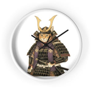 Op de afbeelding: Ronde wandklok met een rode kat afgebeeld als samoerai. De kat draagt traditionele harnas en helm en houdt een katana vast. De klok heeft zwarte wijzers en een witte achtergrond.