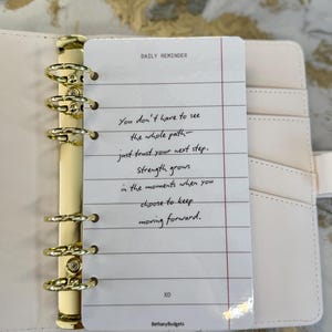 May include: A light pink planner with gold detailing, open to a lined paper insert. The insert features the text "DAILY REMINDER" and a motivational quote. The planner rests on a marble-effect surface.