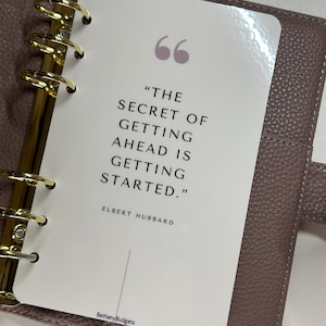 May include: A white planner page with a quote by Elbert Hubbard that reads "The secret of getting ahead is getting started." The quote is surrounded by quotation marks and the author's name is printed below the quote. The planner page is in a brown leather planner.