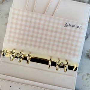 May include: Open, light pink binder with gold-colored rings and a clear pocket labeled "Groceries." The pocket has a tan and white gingham pattern. The binder is open, revealing card slots and a ribbon closure.