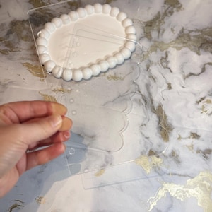 May include: Clear acrylic templates with scalloped edges and a white oval base with a beaded border. The templates have holes for attaching to the base. The background is a marble pattern with gold accents.