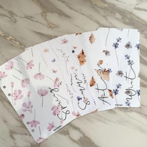 May include: Four white planner dividers with floral designs and seasonal text. The dividers are labelled "Spring," "Summer," "Autumn," and "Winter" in a handwritten font. Each divider features different floral and leaf illustrations in pink, brown, and blue.