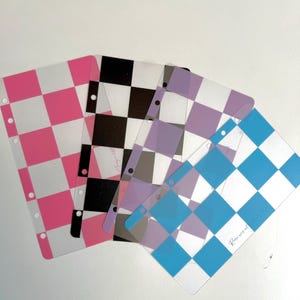 May include: Four rectangular planner dividers with a chequered pattern in pink, black, purple, and blue. Each divider is made of a semi-transparent material and features a vertical row of punched holes along one side for binder insertion.