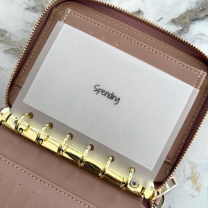 May include: A dusty rose zipped binder featuring a gold-toned six-ring metal mechanism. Inside, a translucent frosted plastic divider is inserted into the rings, displaying the word Spending printed in a simple black sans-serif font.