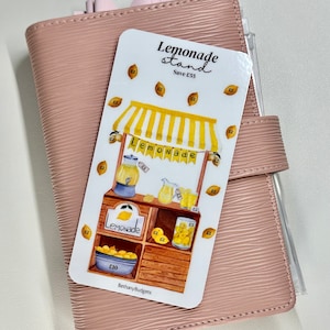 May include: A white printable sticker with a watercolour illustration of a lemonade stand. The stand has a yellow awning with the word "Lemonade" written on it. There are lemons, pitchers, and glasses of lemonade on the stand. The sticker says "Lemonade stand" and "Save £55" at the top and "Lemonade £10" at the bottom. The sticker also has the text "BethanyBudgets" at the bottom.