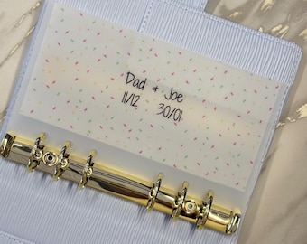 May include: A light blue six-ring binder with a textured finish and gold-tone metal rings. Inside, a translucent vellum sheet features a colorful sprinkle pattern and the printed text "Dad & Joe 11/12 30/01" in a simple black sans-serif font.