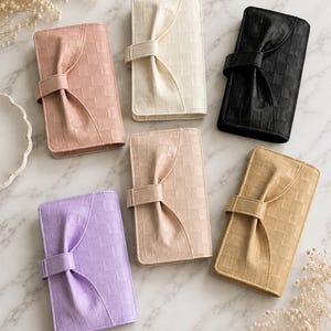 May include: Six rectangular wallets in pink, cream, black, lavender, beige, and tan. Each wallet features a textured checkerboard pattern and a central vertical strap with a decorative bow detail on the front cover, secured with a side snap closure.