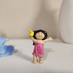 Puede incluir: Una figura de muñeca Cabbage Patch Kid con un traje de baño de lunares rosa y blanco y sosteniendo un pato de goma amarillo.