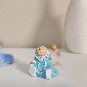 May include: A small figurine of a baby wearing a blue dress with white trim and a bonnet. The baby is holding a rattle and a bottle. The figurine is on a white surface.