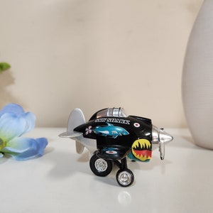 Op de afbeelding: Een zwart en zilveren speelgoedvliegtuig met een blauwe haai en de woorden "Sky Shark" aan de zijkant. Het vliegtuig heeft een rood en geel ontwerp op de staart.