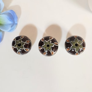 Könnte beinhalten: Drei braune Keramikperlen mit einem weißen und grünen Blumenmuster. Die Perlen haben ein Loch in der Mitte zum Aufziehen.