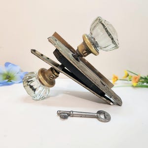 Könnte beinhalten: Antikes Türbeschlagsset mit zwei Türknöpfen aus klarem Glas mit Messingakzenten, einer Metallplatte und einem silbernen Schlüssel. Das Beschlagsset ist gealtert und weist Gebrauchsspuren auf.