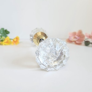 May include: Two clear, faceted glass door knobs with brass hardware. One knob is in focus, displaying a detailed, floral-like pattern. The other knob is slightly out of focus. Decorative flowers in shades of yellow and pink are in the background.