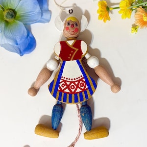 May include: A wooden pull-string doll dressed in a traditional Dutch outfit. The doll features a red vest, white apron with a geometric design, and blue skirt. The doll's arms and legs are jointed, and it has a painted face and a white bonnet.