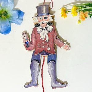 May include: A vintage wooden pull-string toy depicting a gentleman in a red coat, blue trousers, and tall purple boots. He wears a top hat, an eye patch, and holds a miniature nutcracker. A red string hangs from the toy.