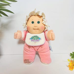 May include: A vintage Cabbage Patch Kids doll, wearing a pink onesie and a matching bonnet. The doll has a round face with rosy cheeks and green eyes. It has a bib with the text "Babyland". The doll's hands are raised in a thumbs-up gesture.
