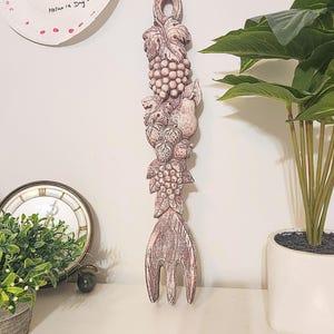 Peut inclure: Fourchette décorative en bois sculpté avec un motif de fruits et de feuilles. La fourchette est peinte dans des tons de rose et de blanc, avec une boucle en haut pour l'accrocher. Le motif comprend des raisins, des poires et d'autres fruits. La fourchette mesure environ 38 cm de long.