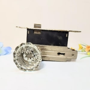 Peut inclure: Un bouton de porte en verre transparent et facetté avec une serrure de porte en métal vintage. Le bouton de porte a un design circulaire en forme de fleur. La serrure est rectangulaire et possède un trou de serrure. Des fleurs décoratives sont en arrière-plan.