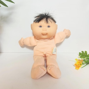 Può includere: Una bambola vintage Cabbage Patch Kid con corpo color pesca e capelli neri. La bambola ha gli occhi blu e indossa una tutina color pesca. La bambola è seduta con le braccia tese.