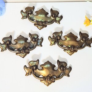 May include: Four antique brass drawer pulls with ornate, symmetrical designs. Each pull features a curved handle and decorative flourishes, with a warm, aged patina. These vintage hardware pieces are ideal for furniture restoration projects.