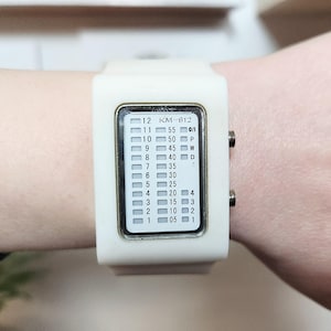 May include: A white digital watch with a rectangular face is shown on a wrist. The watch face has a grid of small rectangles and displays the text "KM-812". Two small black buttons are on the side of the watch.