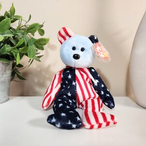 May include: A blue and white beanie baby bear wearing a red, white, and blue American flag pattern. The bear has a red and white striped ear and a blue nose. The bear is sitting on a white surface. The bear has a Ty tag attached to its ear.