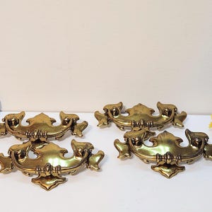 May include: Four antique brass drawer pulls with a decorative design. The pulls have a curved shape with ornate detailing and a warm, golden finish. Perfect for adding a vintage touch to furniture.