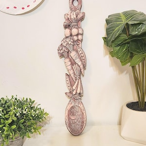 May include: A wooden spoon with a carved design of vegetables, including carrots, peas, and a bunch of radishes. The spoon is painted in a distressed white and pink finish.