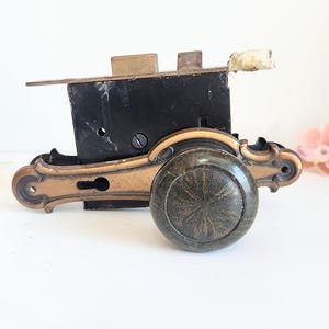 May include: An antique door lock with a bronze finish. The lock features a black metal box, a decorative bronze handle, and a round, textured knob. The design includes ornate details and a keyhole. The hardware is set against a white background.