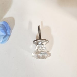 Könnte beinhalten: Ein klarer Glas-Schrankknopf mit einer silbernen Metallbasis und einer Schraube. Der Knopf hat ein facettierendes Design.