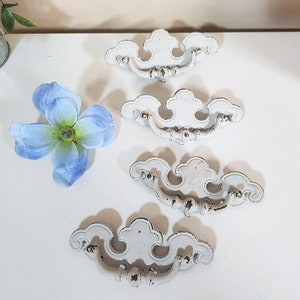 May include: Four white, ornate drawer pulls with a decorative, scalloped design. The pulls have a distressed, vintage look and are perfect for adding a touch of elegance to any furniture piece.