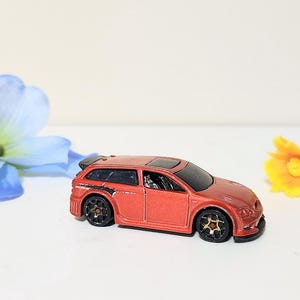 May include: A red toy car with black wheels and a black spoiler. The miniature vehicle is a station wagon style, with a black roof and tinted windows. The car is set against a white background with blue and yellow flowers.